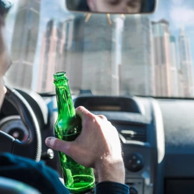 A man holding a beer bottle while driving, with a city skyline visible through the windshield.