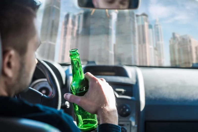 A man holding a beer bottle while driving, with a city skyline visible through the windshield.