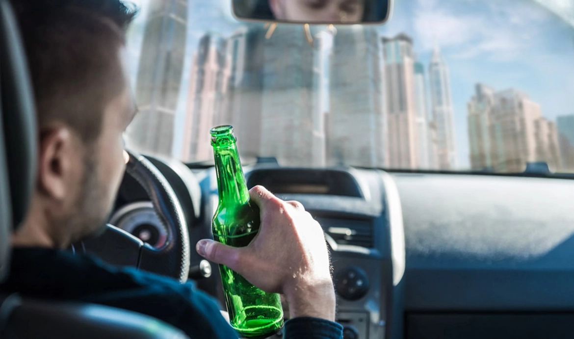 A man holding a beer bottle while driving, with a city skyline visible through the windshield.