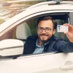 A man sitting in a car holds up his driver's license while smiling.