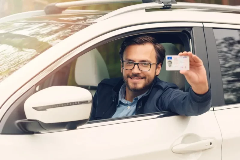 A man sitting in a car holds up his driver's license while smiling.